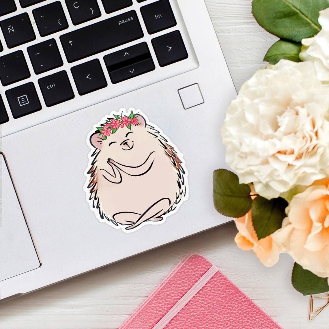 A hedgehog is sitting with a yoga pose and is wearing a pink flower crown. The hedgehog has a happy expression on its face. The sticker is placed next to the mousepad on a laptop on a wooden desk with flowers in view to the right.