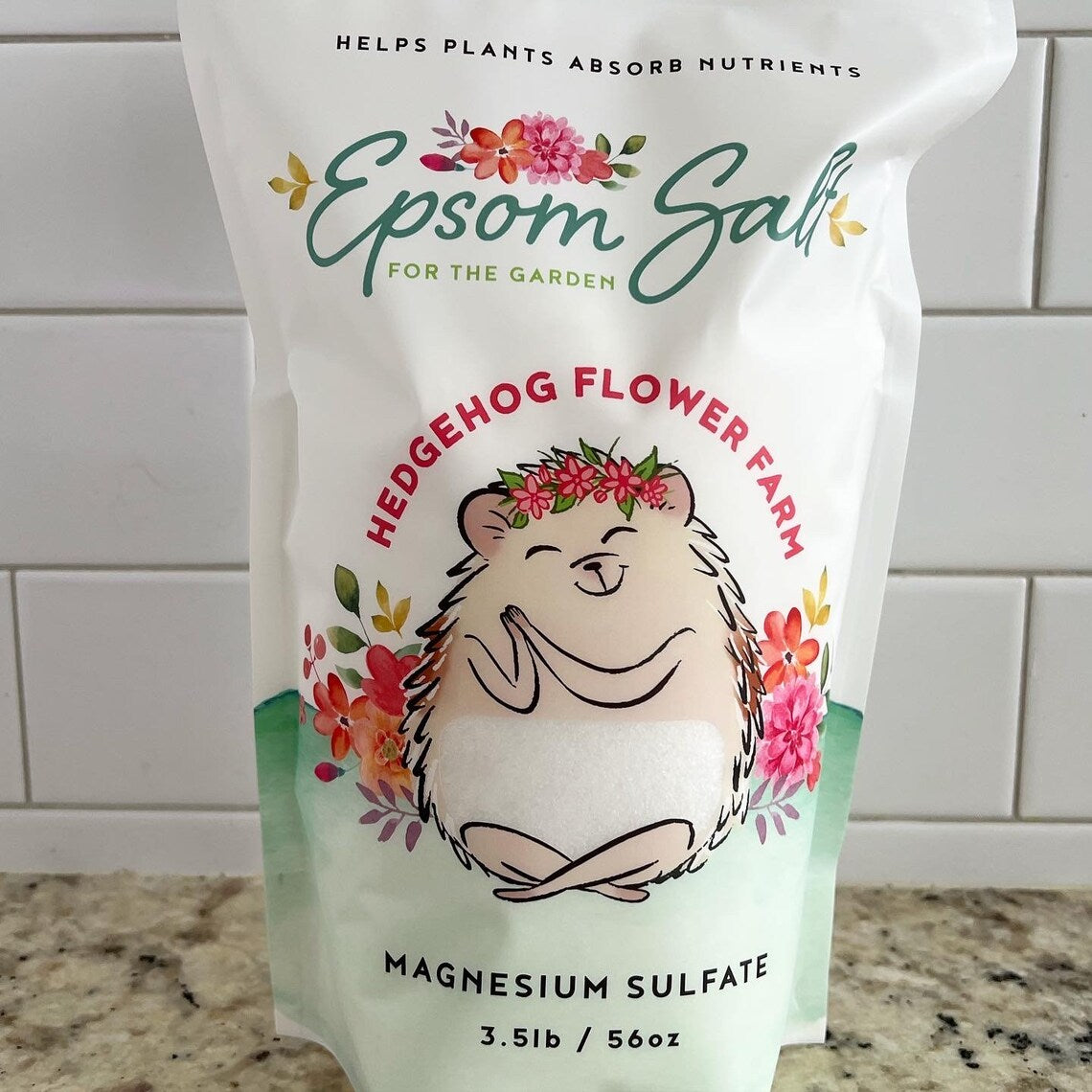 A white bag of Epsom salt for the garden. The bag features a cartoon hedgehog with a flower crown and the words 'Hedgehog Flower Farm' and 'Magnesium Sulfate 3.5lb / 56oz'. Background is white tile wall on a beige and black spotted counter.