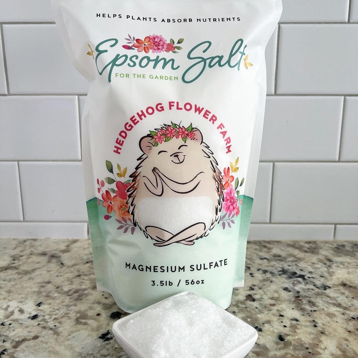A white bag of Epsom salt for the garden. The bag features a cartoon hedgehog with a flower crown and the words 'Hedgehog Flower Farm' and 'Magnesium Sulfate 3.5lb / 56oz'. Background is white tile wall on a beige and black spotted counter with a square bowl of epsom salt shown in front of the bag.