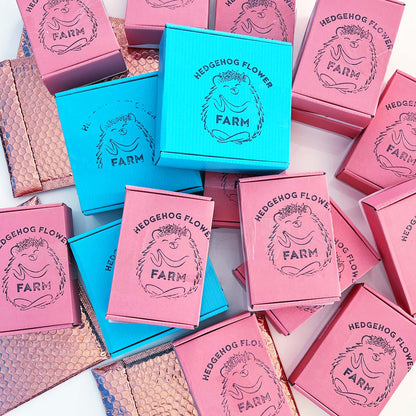 A collection of small, square boxes with a hedgehog design. The boxes are in shades of pink and blue and feature the text 'Hedgehog Flower Farm' with a hedgehog illustration. The boxes are stacked and arranged in a random pattern.
