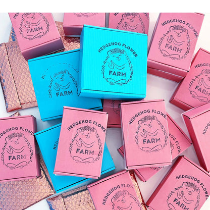 A collection of small, square boxes with a hedgehog design. The boxes are in shades of pink and blue and feature the text 'Hedgehog Flower Farm' with a hedgehog illustration. The boxes are stacked and arranged in a random pattern.
