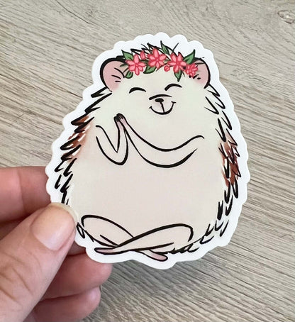 A hedgehog is sitting with a yoga pose and is wearing a pink flower crown. The hedgehog has a happy expression on its face. This is a sticker being held up by someone against a wooden style background. 