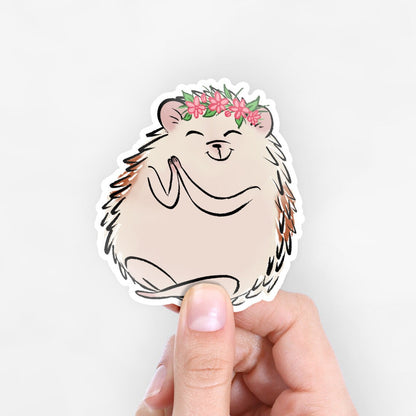 A hedgehog is sitting with a yoga pose and is wearing a pink flower crown. The hedgehog has a happy expression on its face. This is a sticker being held up by someone against a white background. 