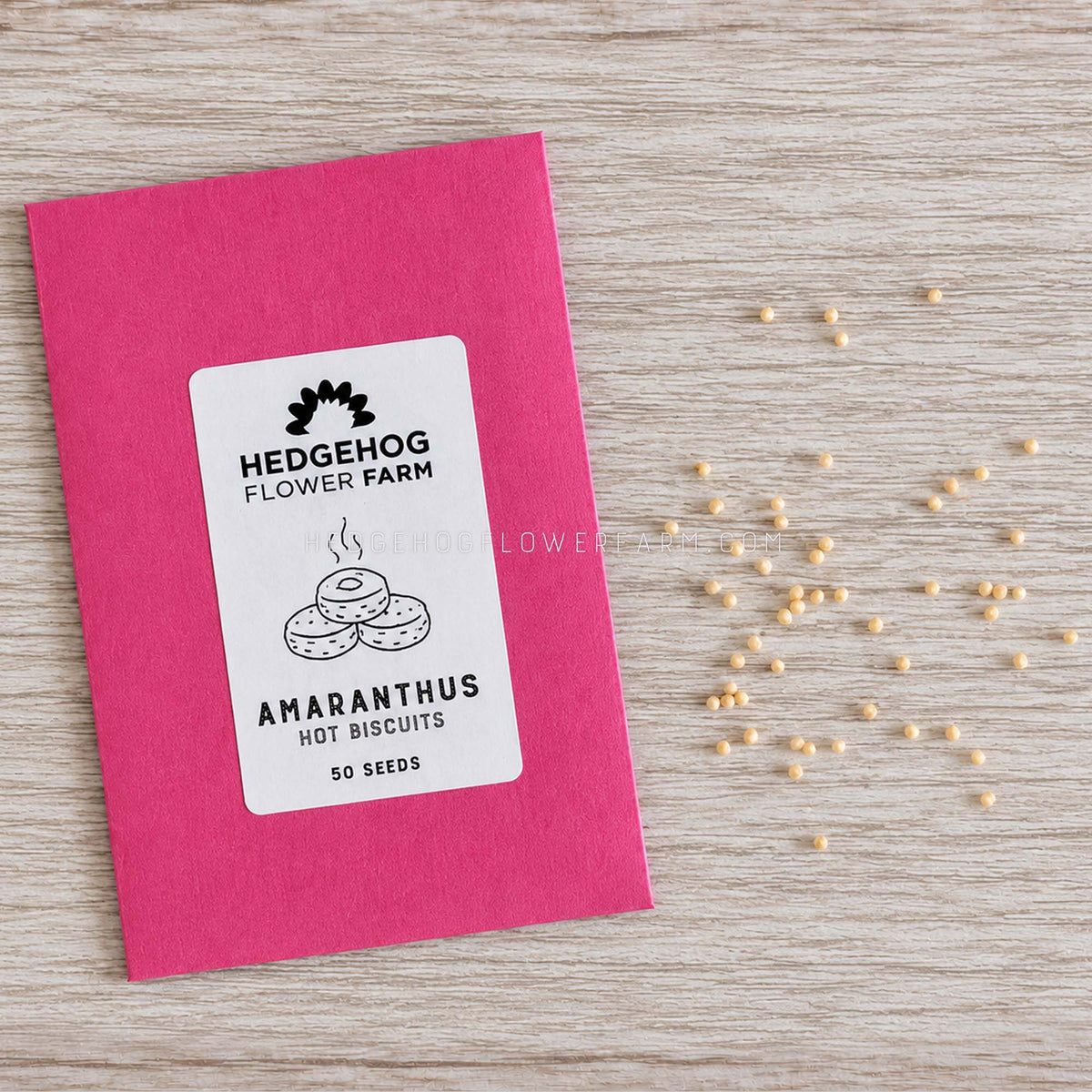 A pink seed packet labeled 'Hedgehog Flower Farm Amaranthus Hot Biscuits' lies on a light wood surface with scattered small beige seeds nearby.