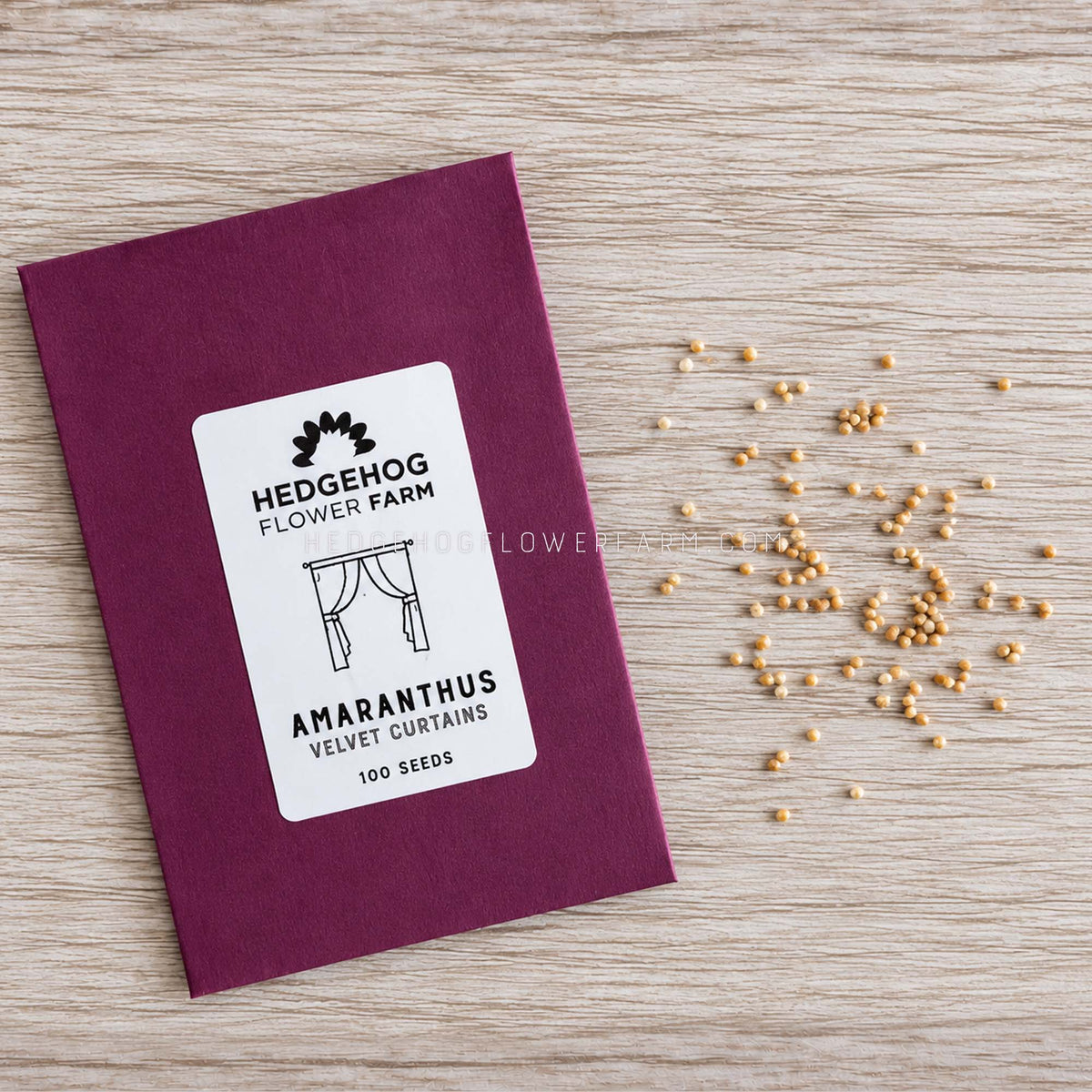 A seed packet for 'Amaranthus Velvet Curtains' by Hedgehog Flower Farm, placed on a light wood grain surface next to scattered golden seeds.