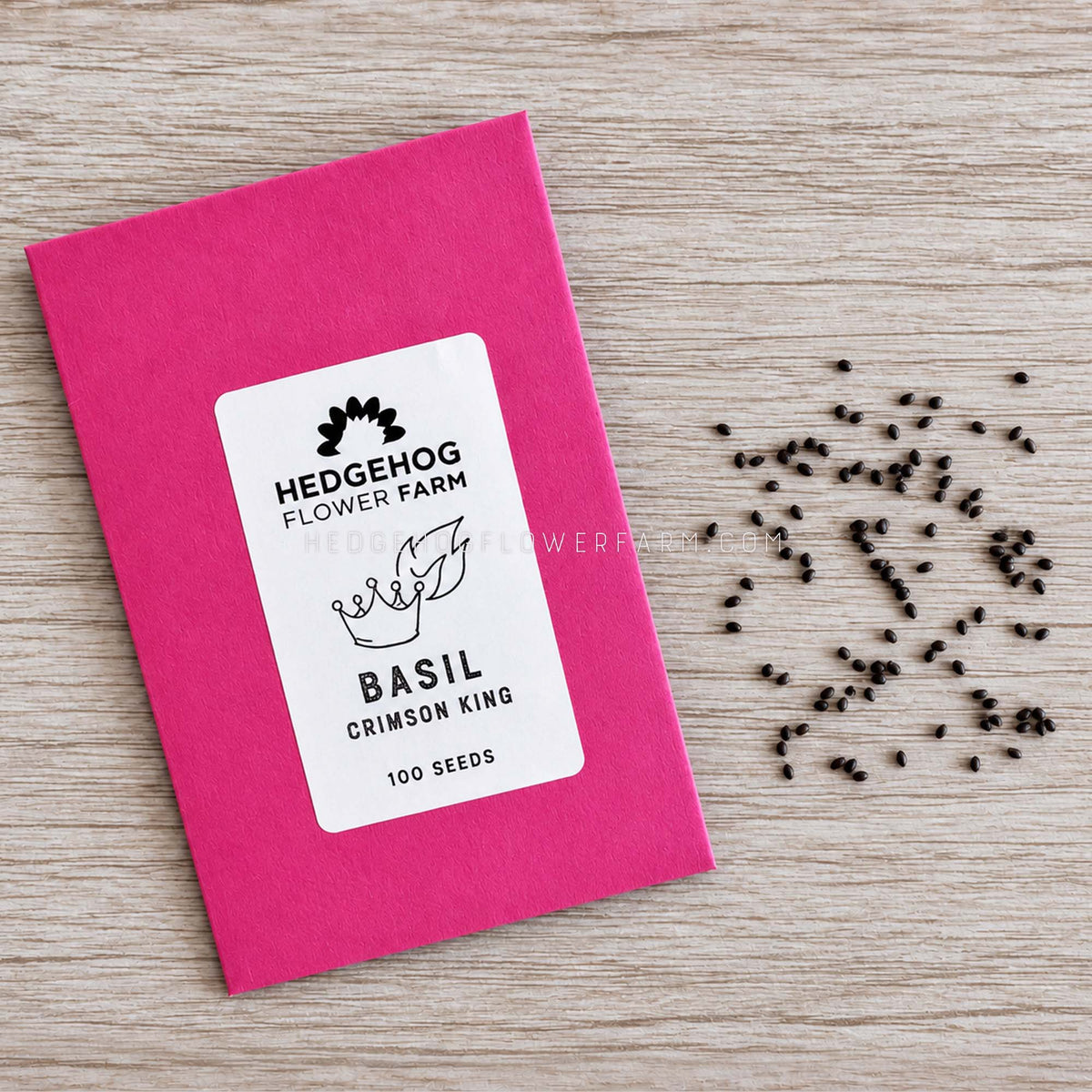 A bright pink seed packet labeled Hedgehog Flower Farm Basil Crimson King, with black seeds scattered on a light wood surface.