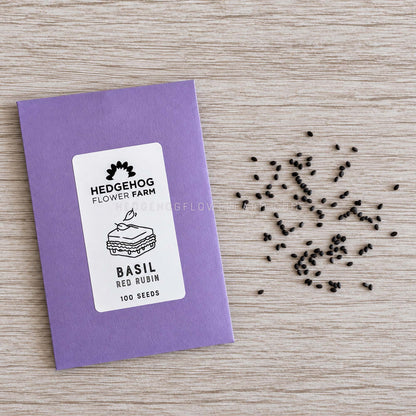 A purple seed packet labeled Hedgehog Flower Farm Basil Red Rubin, placed on a light wood surface with several black seeds scattered beside it.