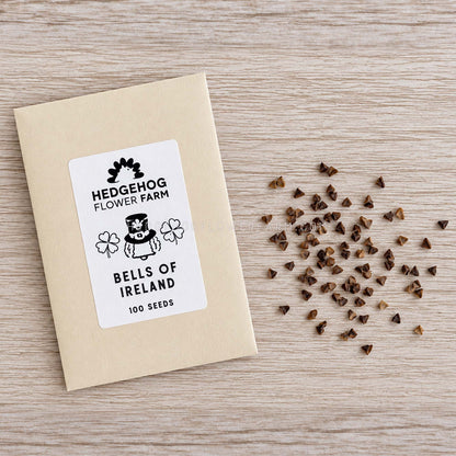 A kraft paper seed packet labeled 'Hedgehog Flower Farm Bells of Ireland 100 Seeds' lies on a light wood surface with scattered brown seeds to the right.