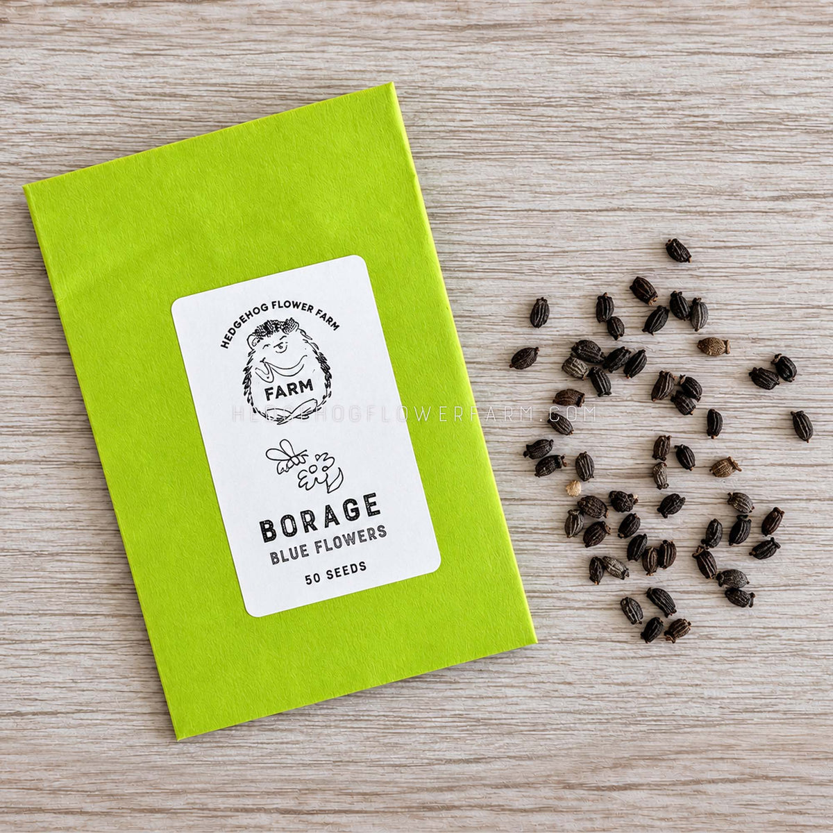 A kraft paper seed packet labeled 'Hedgehog Flower Farm Borage Blue Flowers 50 Seeds' lies on a light wood surface with scattered brown seeds to the right.
