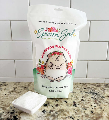 A white bag of Epsom salt for the garden. The bag features a cartoon hedgehog with a flower crown and the words 'Hedgehog Flower Farm' and 'Magnesium Sulfate 3.5lb / 56oz'. Background is white tile wall on a beige and black spotted counter with a square bowl of epsom salt shown in front of the bag. 