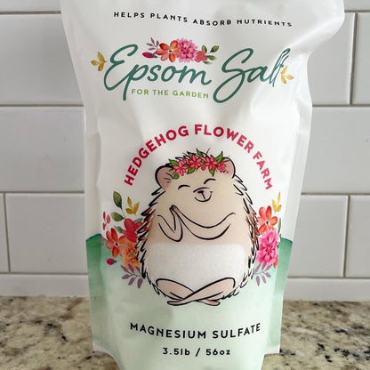 A white bag of Epsom salt for the garden. The bag features a cartoon hedgehog with a flower crown and the words 'Hedgehog Flower Farm' and 'Magnesium Sulfate 3.5lb / 56oz'. Background is white tile wall on a beige and black spotted counter.