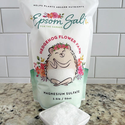A white bag of Epsom salt for the garden. The bag features a cartoon hedgehog with a flower crown and the words 'Hedgehog Flower Farm' and 'Magnesium Sulfate 3.5lb / 56oz'. Background is white tile wall on a beige and black spotted counter with a square bowl of epsom salt shown in front of the bag.